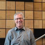 Photo: Thomas Burbacher smiling and standing in front of a wood dedication wall.