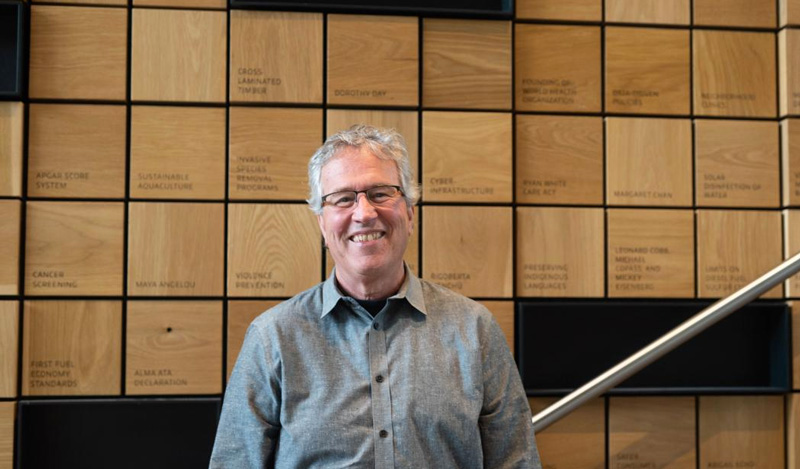 Photo: Thomas Burbacher smiling and standing in front of a wood dedication wall.