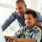 Photo of a father and sitting son in front of a laptop screen reading.