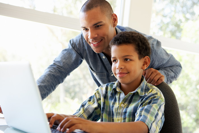 Photo of a father and sitting son in front of a laptop screen reading. 
