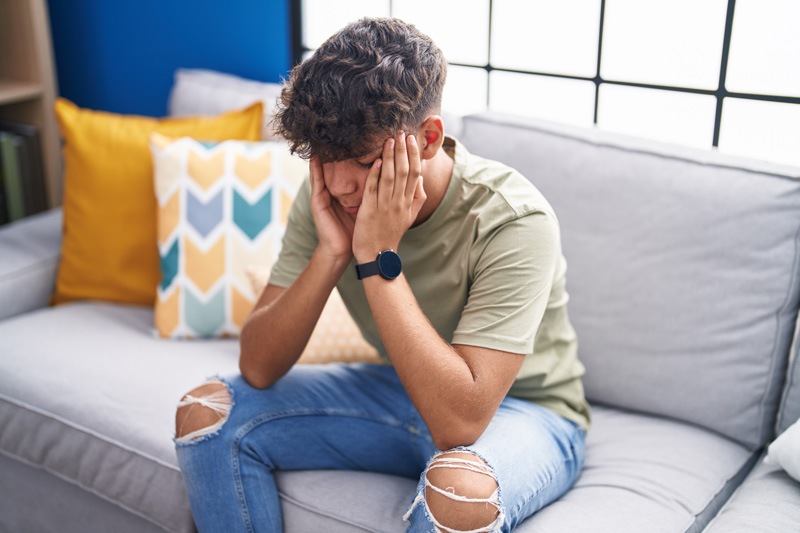 Photo - Young man sitting on a couch with his eyes closed and his head in his hands.