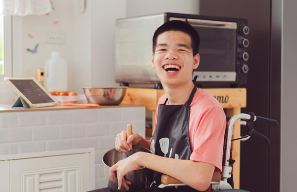 Photo: Smiling young man wearing an apron and in a wheelchair assisting with meal prep in a kitchen. 