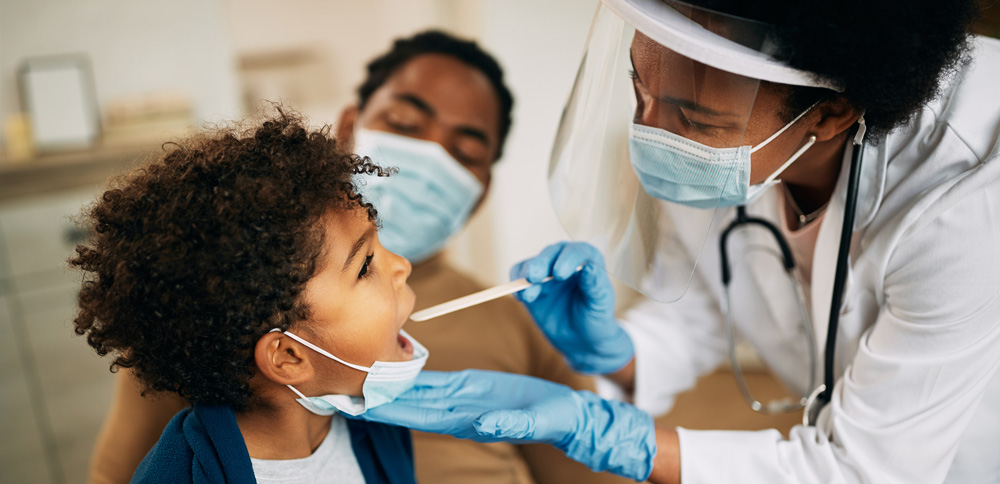 Photo: A clinician, wearing a mask and shield, examining a young patient with their parent in the background wearing a mask.