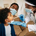 Photo: A clinician, wearing a mask and shield, examining a young patient with their parent in the background.