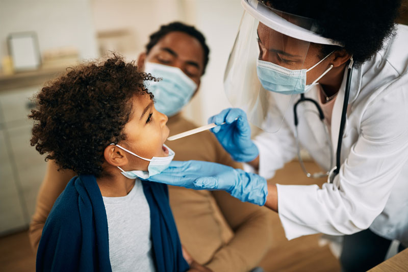 Photo: A clinician, wearing a mask and shield, examining a young patient with their parent in the background.