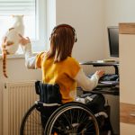 Photo: Young woman wearing headphones, sitting in a wheelchair, petting a cat that is sitting in the window.