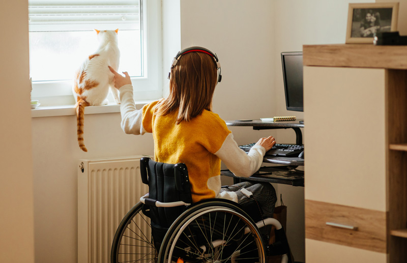 Photo: Young woman wearing headphones, sitting in a wheelchair, petting a cat that is sitting in the window.