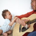 Photo: Infant smiling and holding onto mom while she plays the guitar.