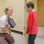 Photo: A smiling child looking at an Augmented Alternative Communication device held by his smiling instructor and another instructor is smiling in the doorway.