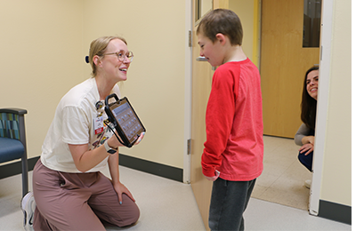 Photo: A smiling child looking at an Augmented Alternative Communication device held by his smiling instructor and another instructor is smiling in the doorway.