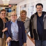 Photo: Four people standing and smiling in a hallway. One person has a cane.