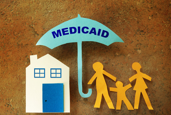 Image: Paper cut-outs of a home and family covered by an umbrella branded with 'Medicaid.'