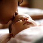 Link to the Duncan Conference online registration. Photo: Low light photo of a mother kissing a sleeping baby on the forehead.