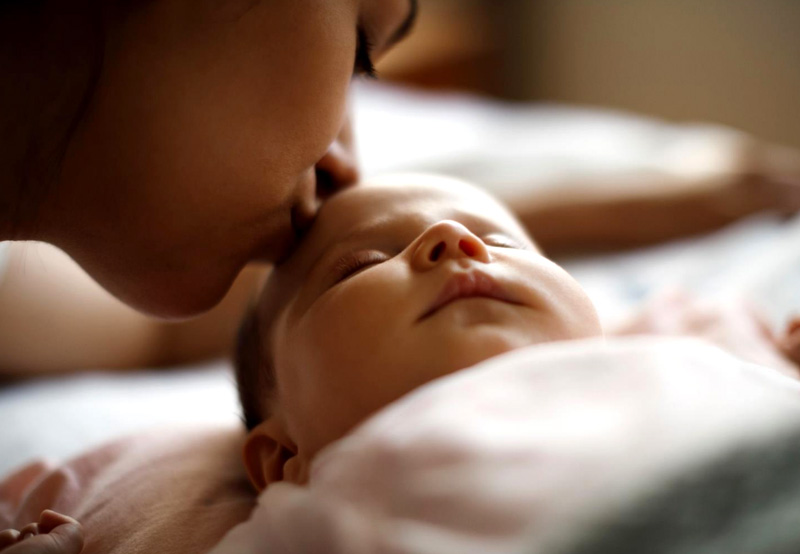 Photo: Low light photo of a mother kissing a sleeping baby on the forehead.