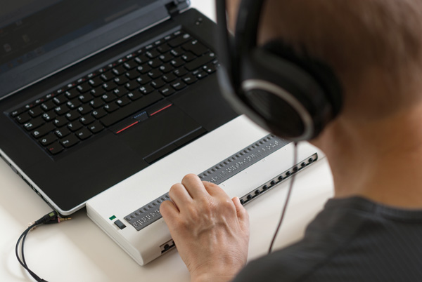 Photo: Blind person working on computer with braille display and screen.