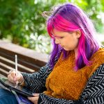 Photo: Young adult drawing while sitting on a park bench.